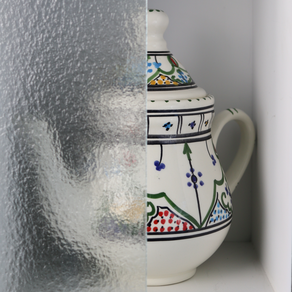 a cabinet glass display with glass covering one side of a teapot, the glass has a rough texture and obscures the pattern but not the shape of the teapot